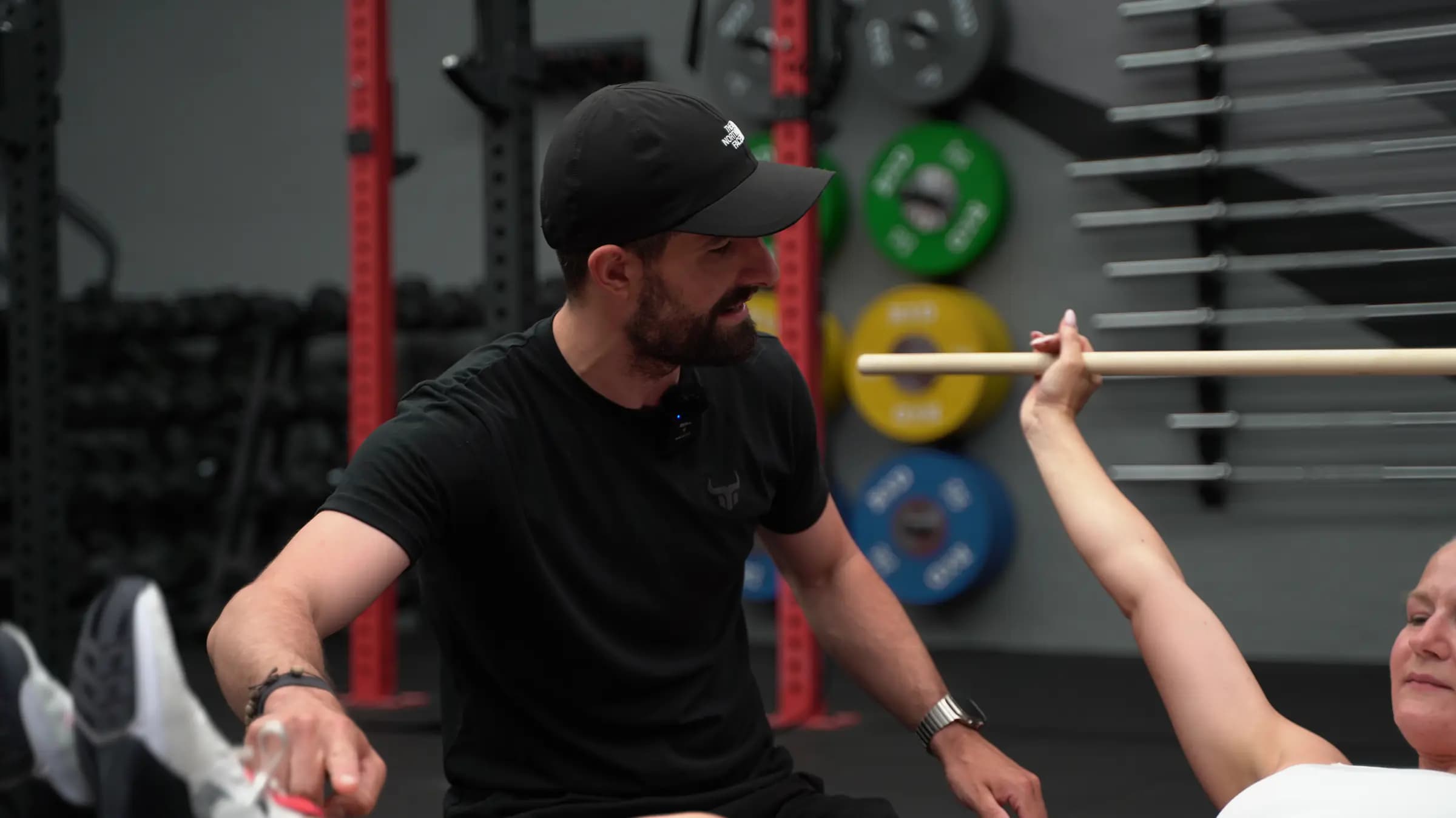 Trainer coacht eine Teilnehmerin am Boden bei einer Überkopf-Übung mit dem Holzstab im Iron Gym Viersen
