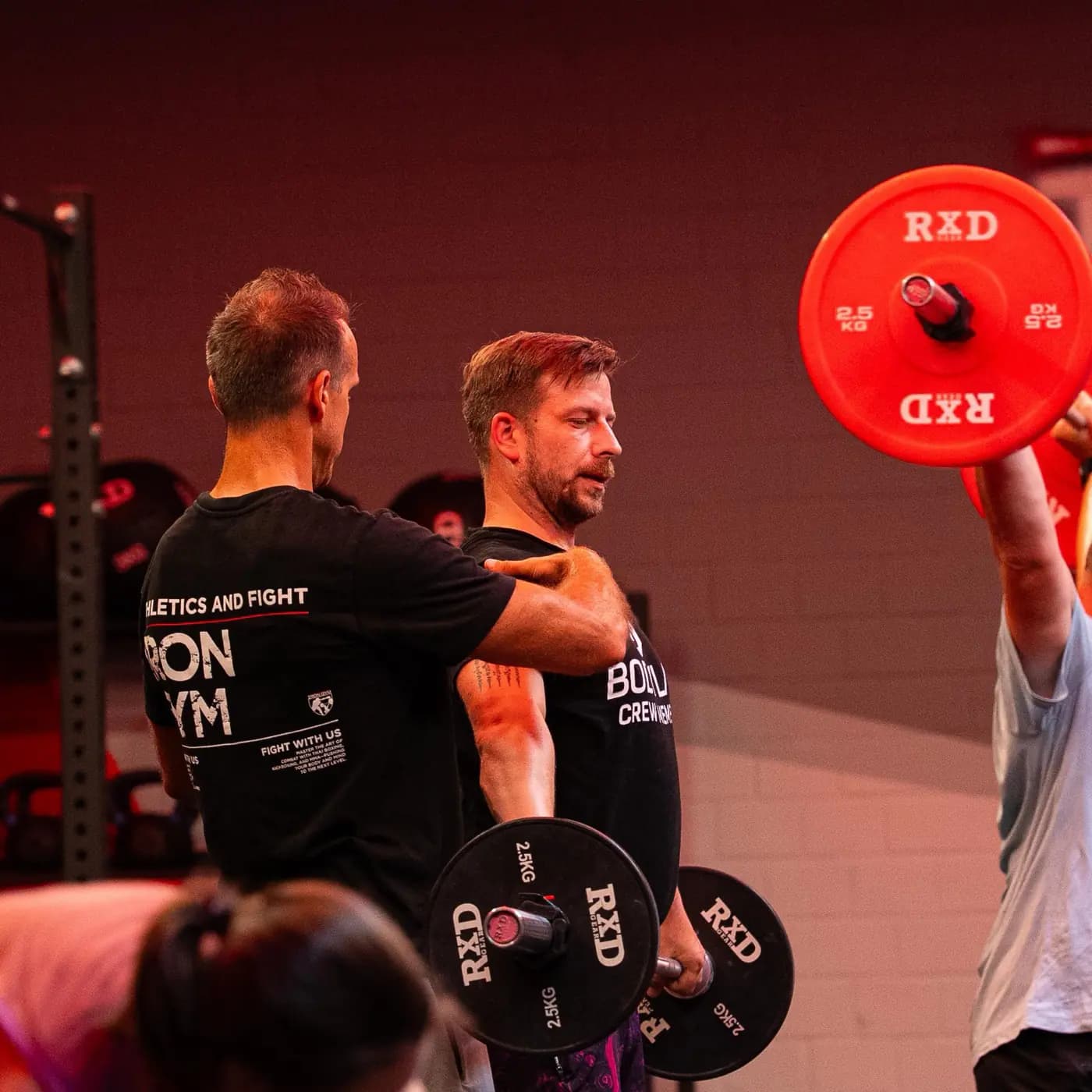 Trainer im Iron-Gym-Shirt korrigiert die Schulterposition an der Langhantel, rechts drückt ein Teilnehmer die Hantel über Kopf