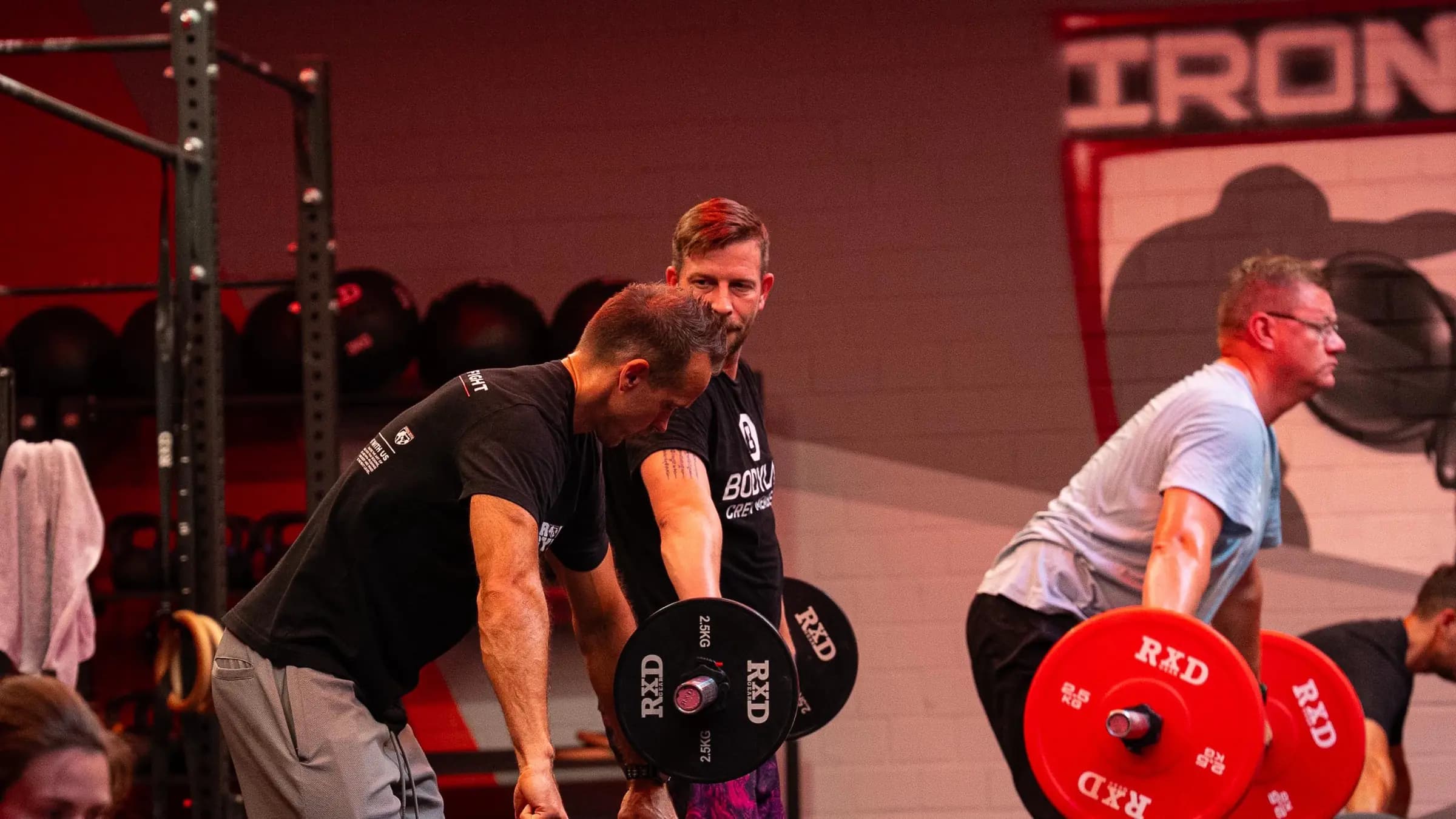 Trainer betreut einen Teilnehmer an der Langhantel im Iron Athletics Kurs, rechts zieht ein zweiter seine Hantel mit roten Scheiben, Iron Gym Viersen