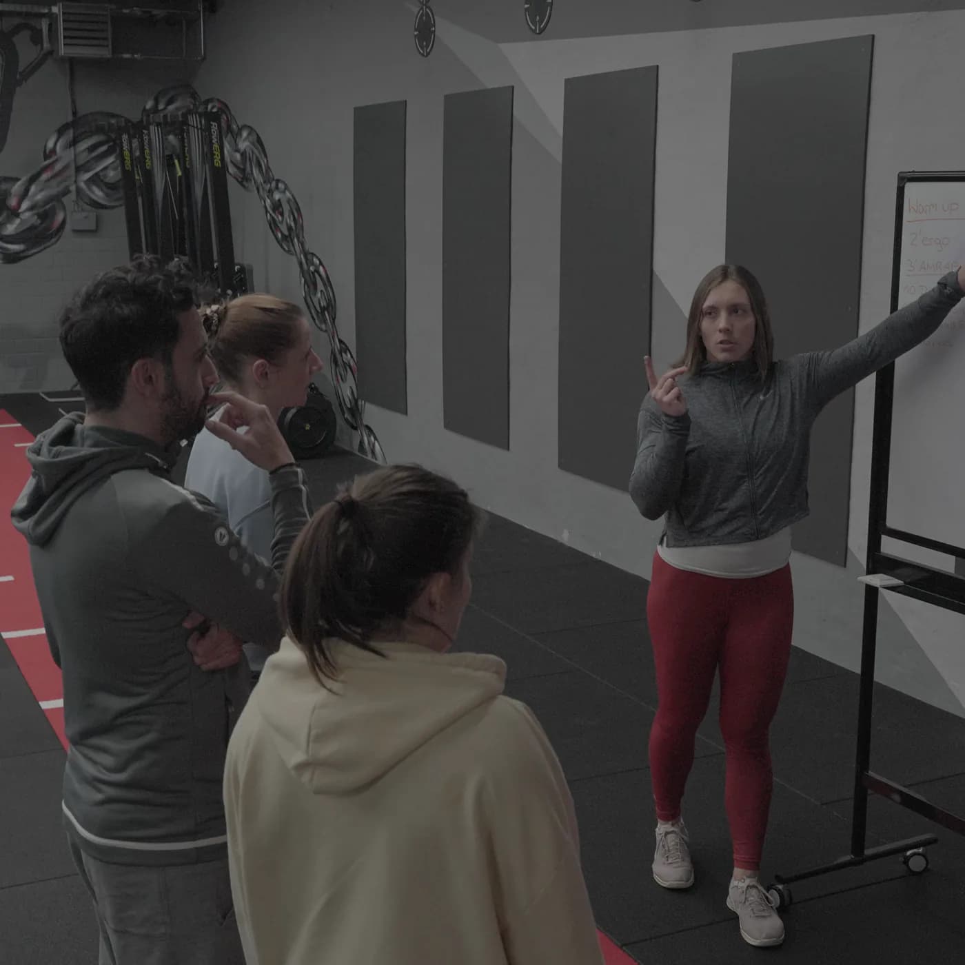 Trainerin erklärt das Workout of the Day am Whiteboard, vier Teilnehmer hören zu