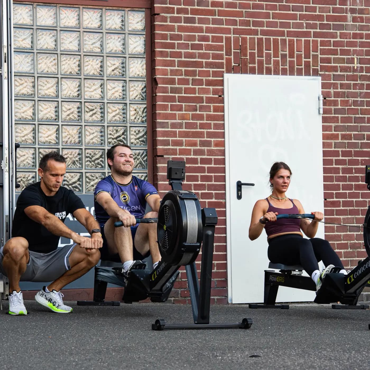 Zwei Sportler ziehen auf Concept2 Ruderergometern vor dem Iron Gym Viersen, der Trainer hockt daneben und beobachtet den Zug