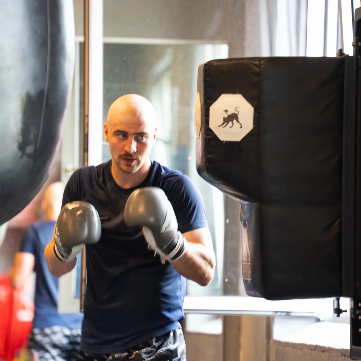 Boxer steht mit Handschuhen in der Grundstellung am Sandsack, rechts das Schlagpolster an der Wand