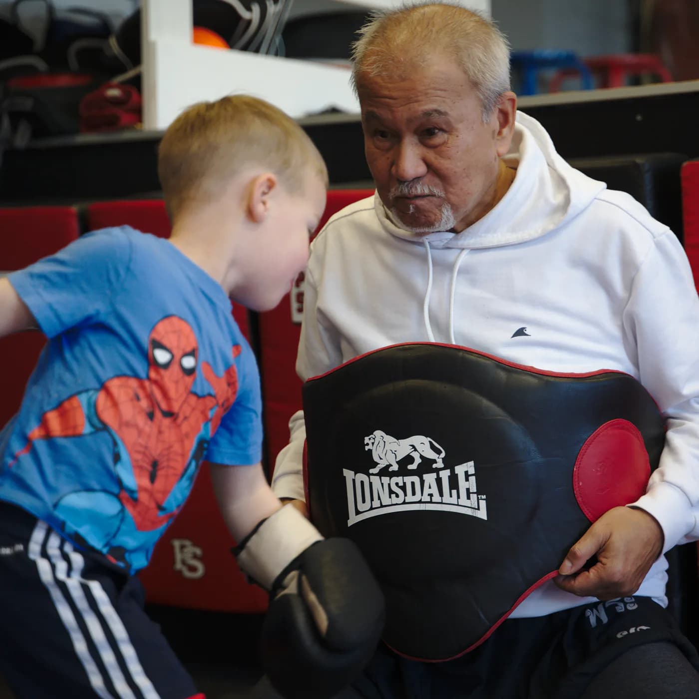 Bambini-Training im Iron Gym: Ein Vorschulkind schlägt mit Boxhandschuhen auf das Bauchpolster des knienden Trainers
