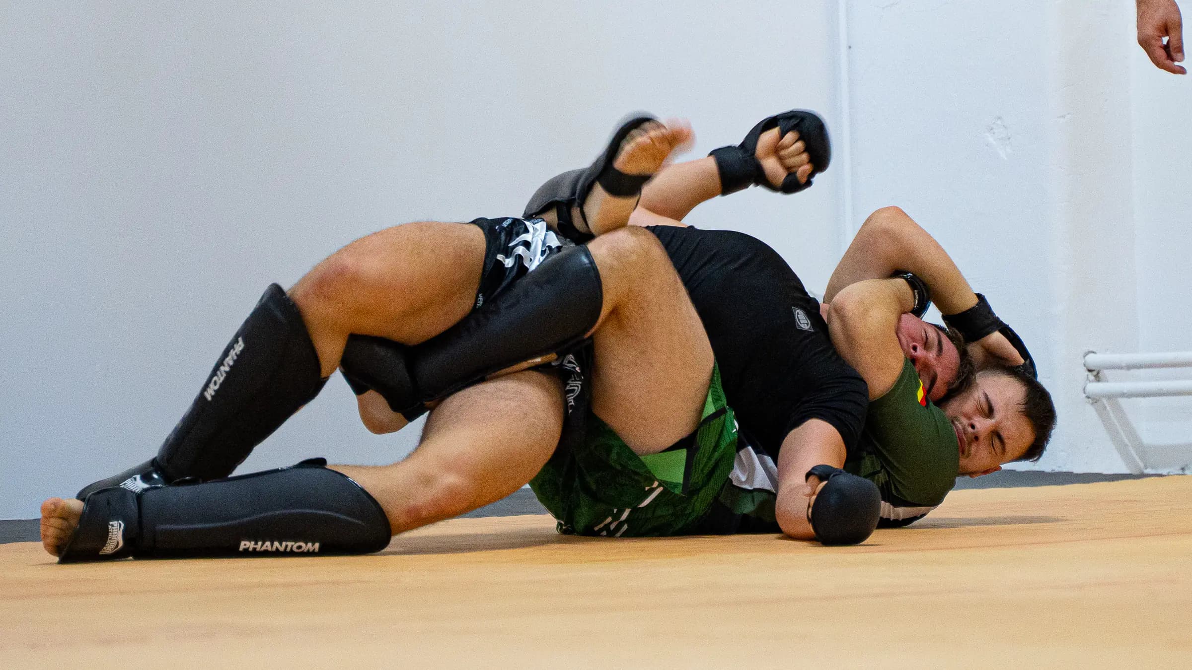 Zwei Sportler im Bodenkampf mit offenen MMA-Handschuhen und Schienbeinschonern im Iron Gym